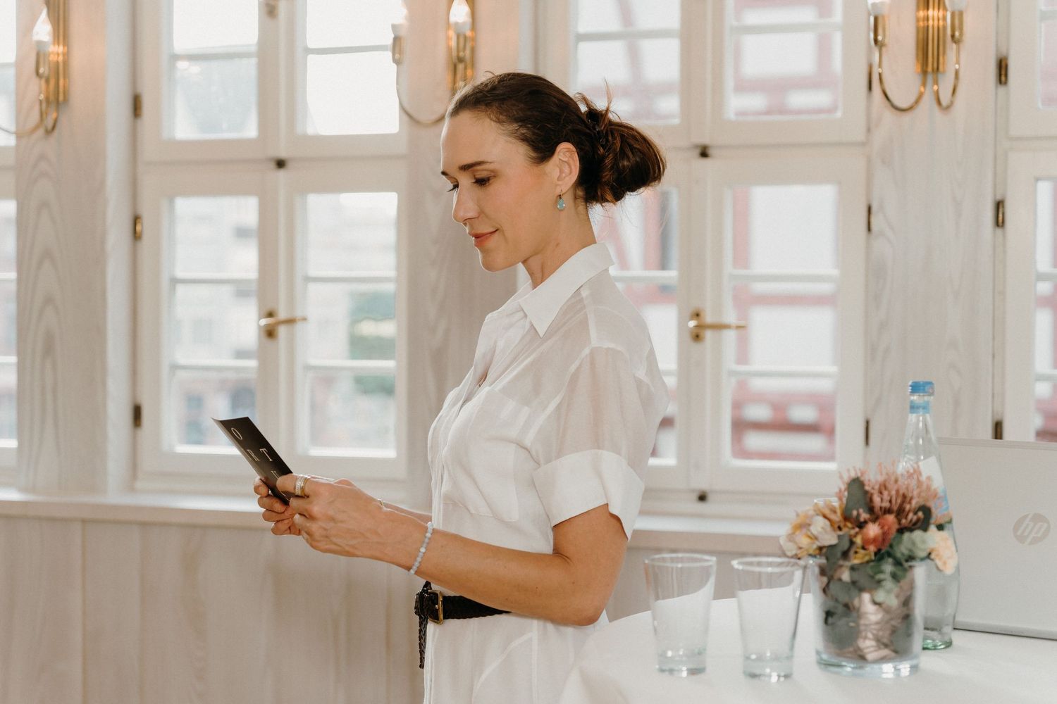 Woman in white shirt checking smartphone near bar with glasses and flowers in elegant interior.