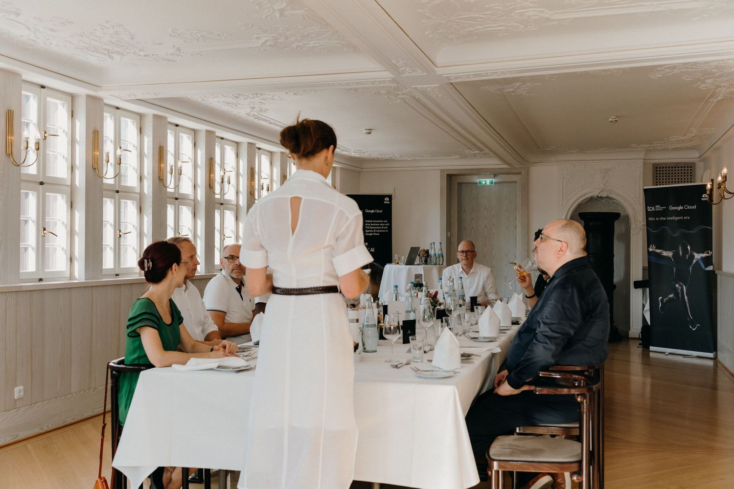 Elegant restaurant dining scene with guests seated at white-clothed tables while a server in white dress attends them.