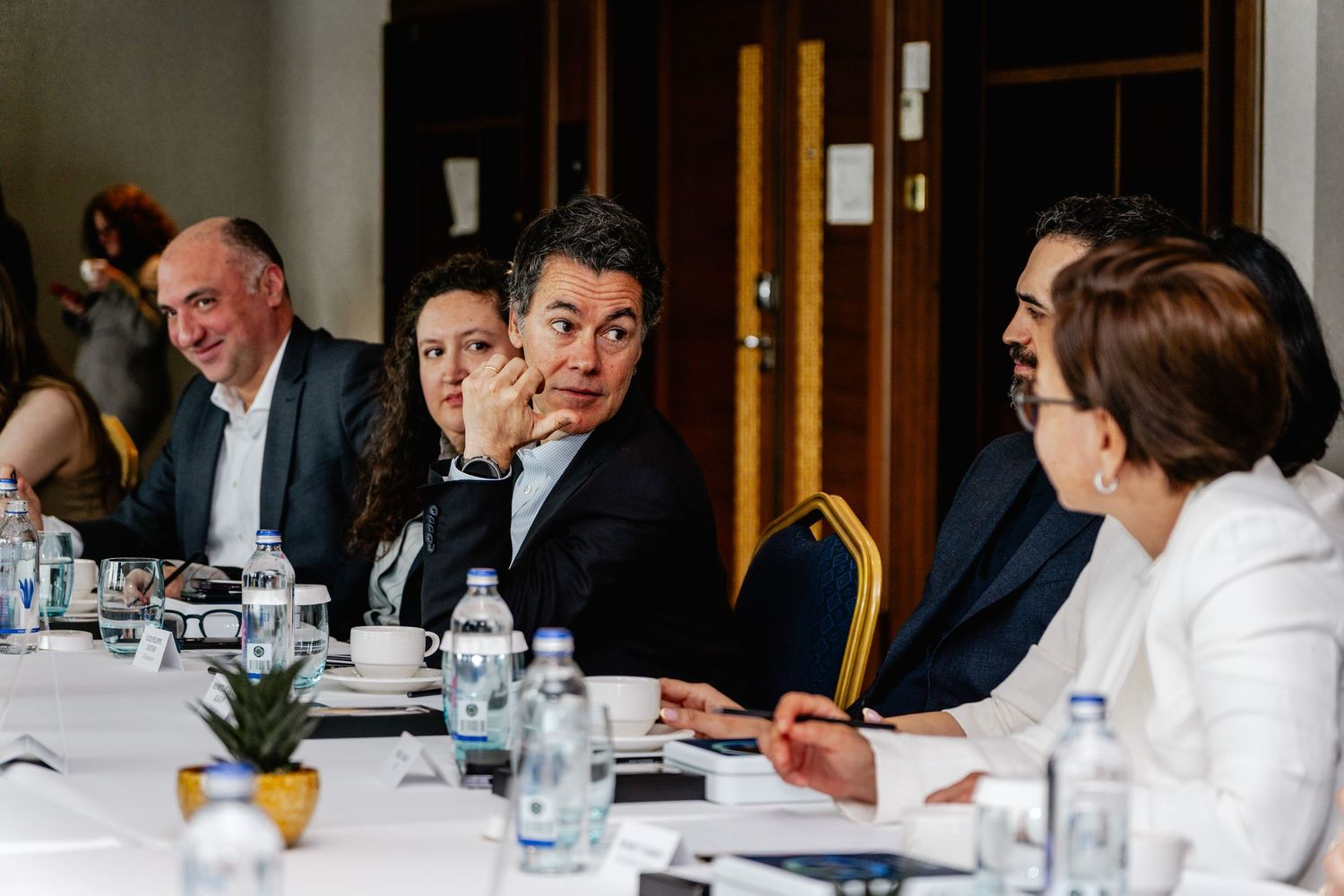 Business professionals engaged in discussion at a corporate meeting table with water bottles and documents.
