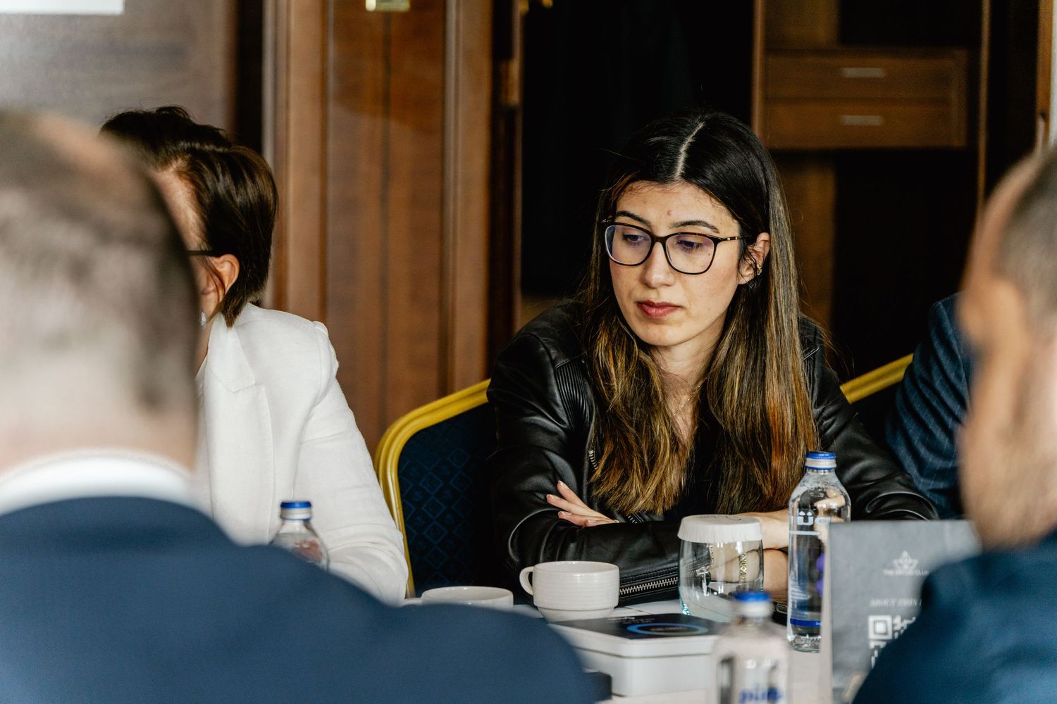Young woman with glasses in a black jacket speaking at a professional business meeting conference table.