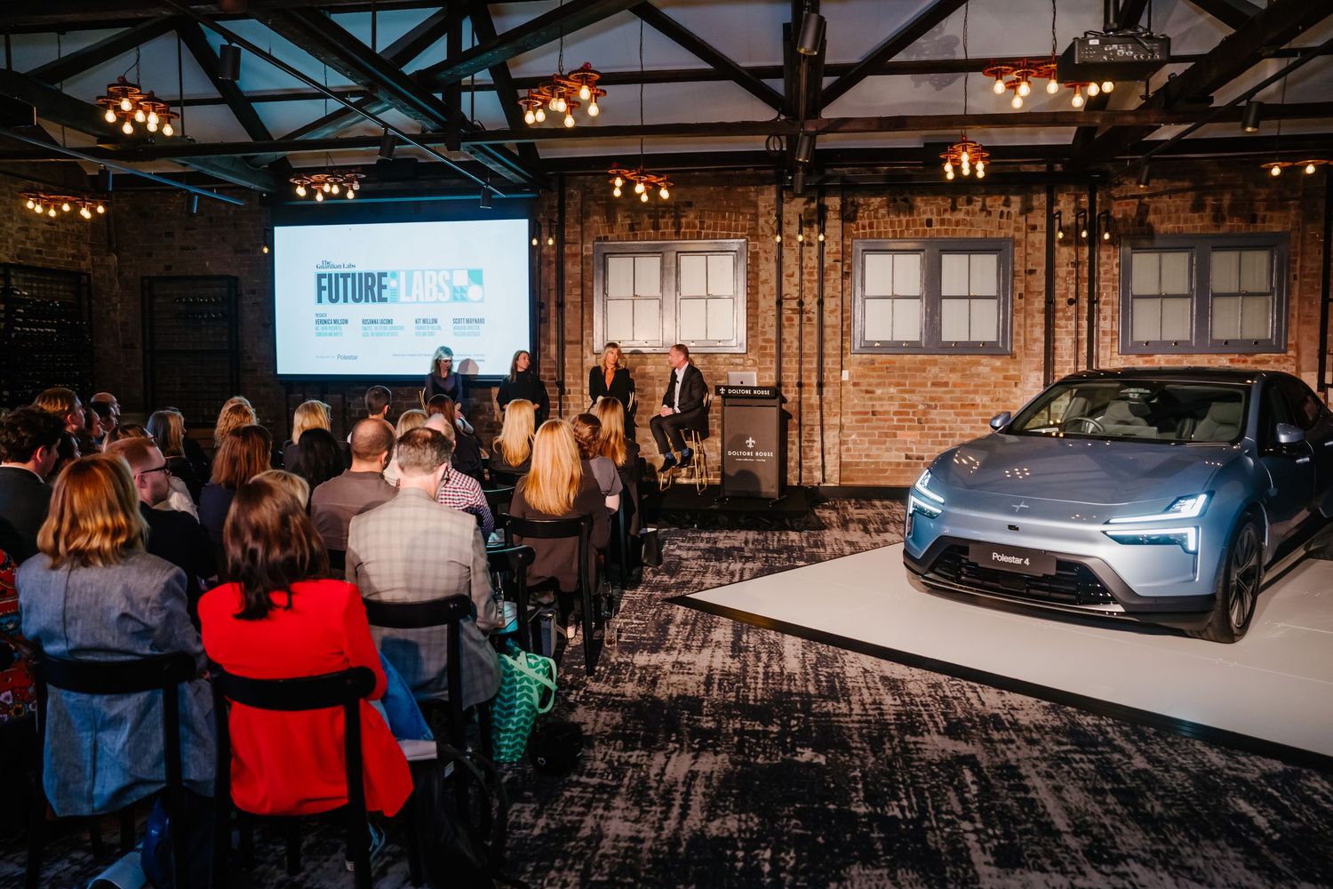 An audience watches a presentation of a silver electric vehicle in an industrial venue with exposed brick and hanging lights.