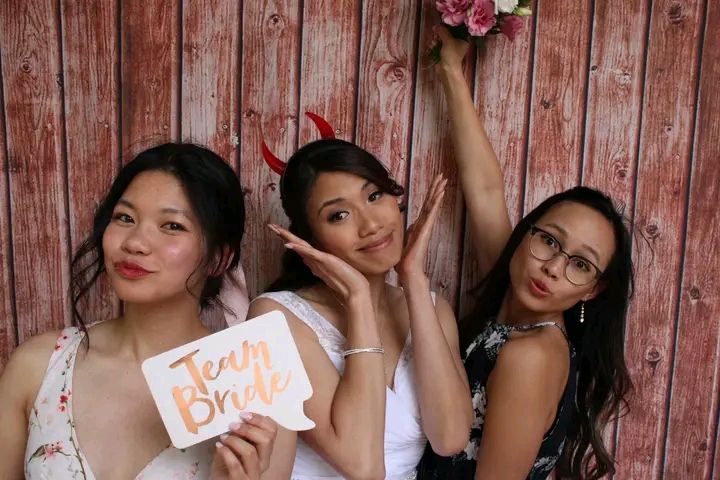 A group of people pose playfully with photo booth props against a rustic wooden wall background.