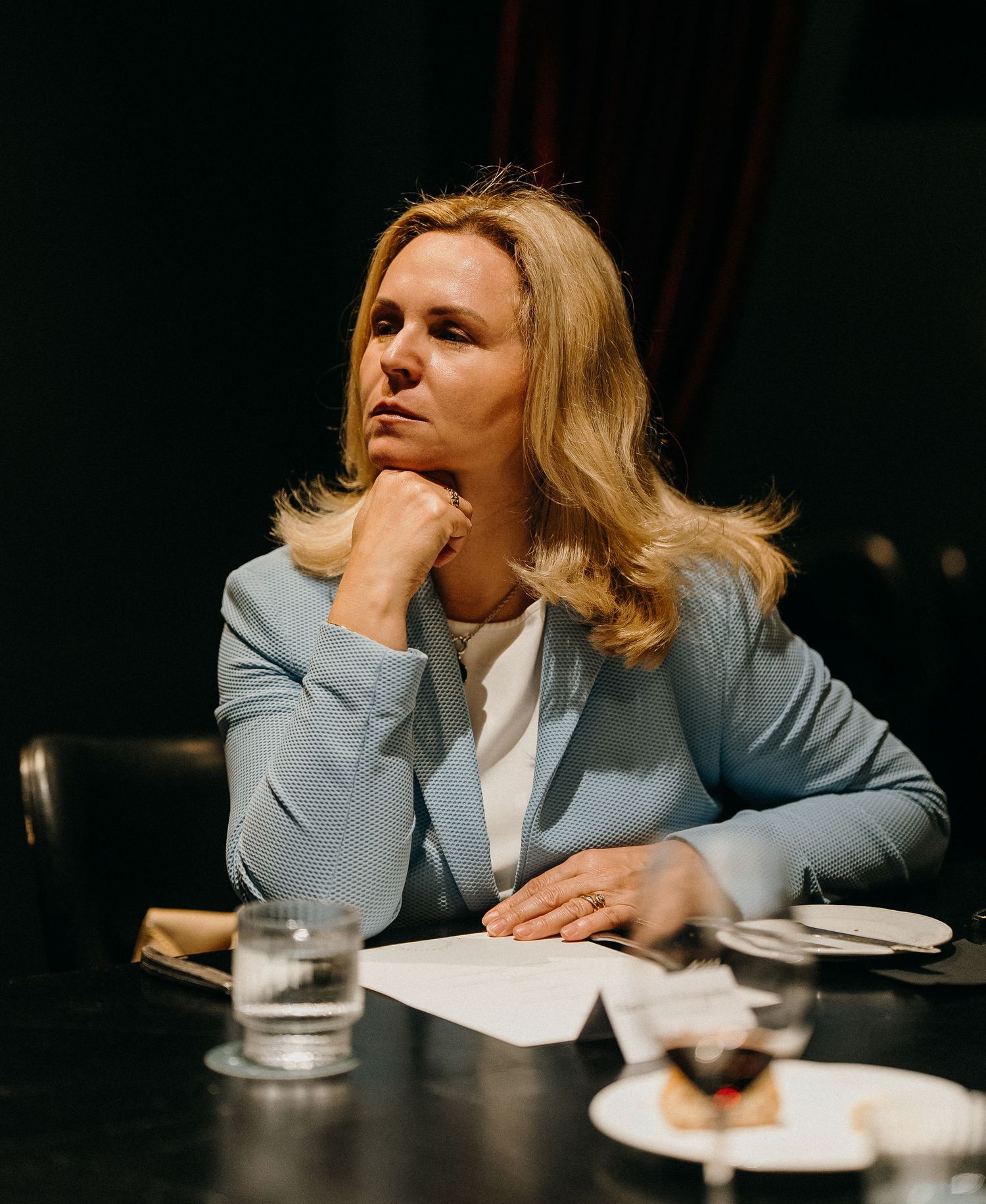 Person in light gray blazer sitting at table with glass of water and dessert plate in dark moody setting.