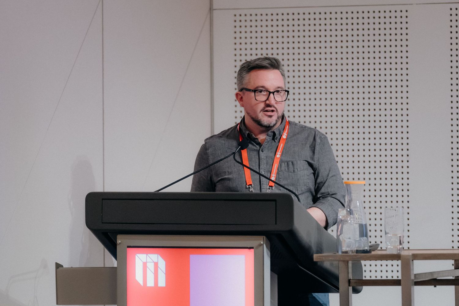 A speaker presents from behind a podium with a red logo during a professional conference presentation.