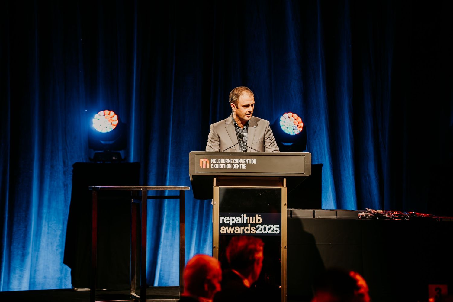 A speaker stands at a podium on stage with blue curtains and lighting during a presentation.