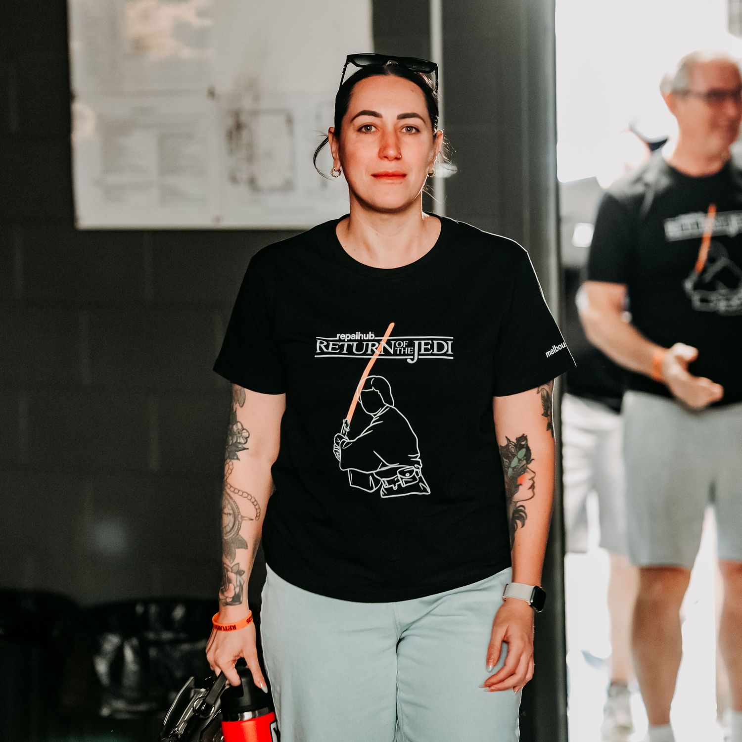 Person in black t-shirt with tattoos and light pants stands indoors while holding camera equipment.