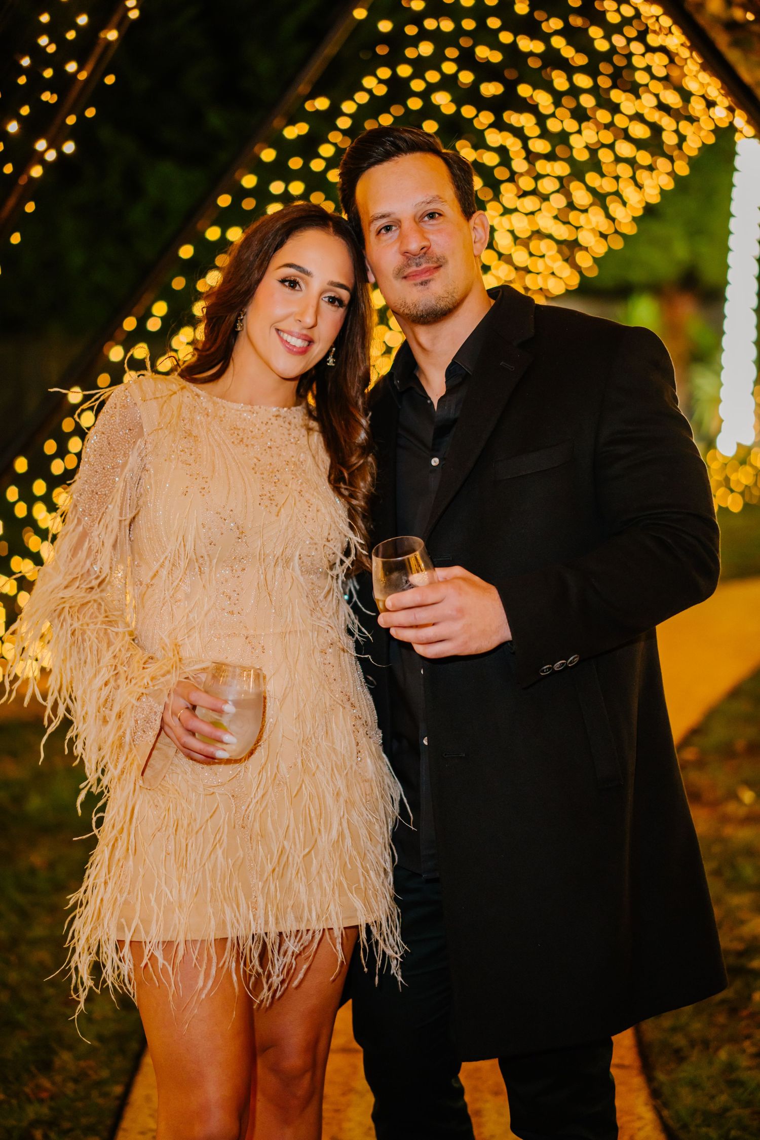 Couple poses together at night against backdrop of twinkling golden lights in romantic outdoor setting.