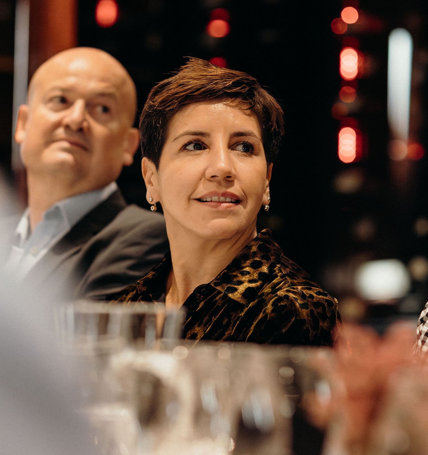 A person in a leopard print top smiles warmly at a formal dinner event with soft lighting and wine glasses visible.