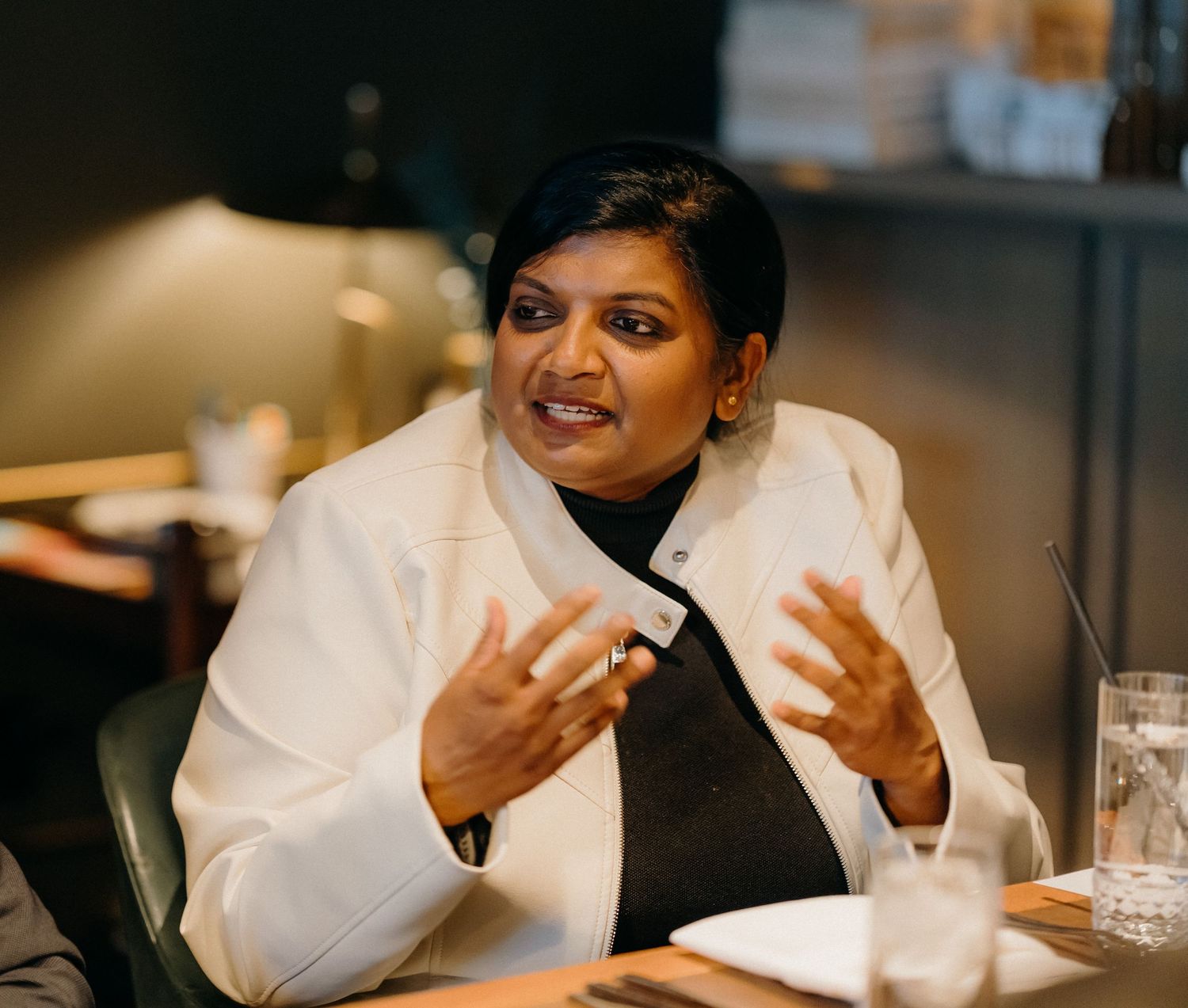 Chef in white coat gestures while engaged in animated conversation at restaurant table.