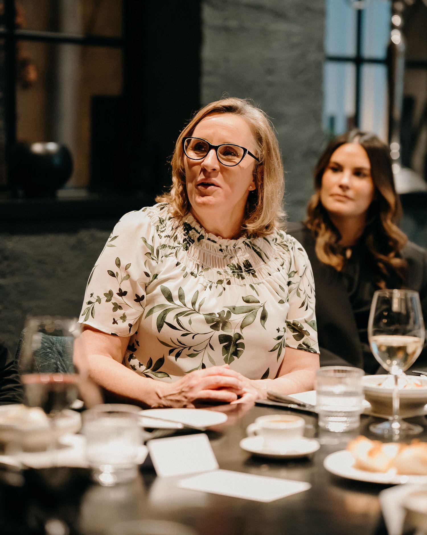 Person in glasses and floral shirt sits at formal dining table with wine glasses and place settings.