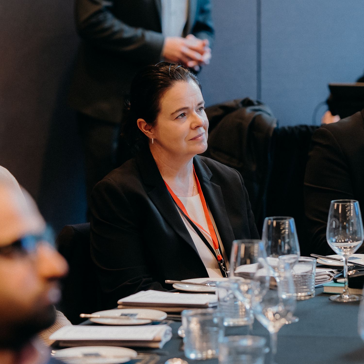 Business professional in a black blazer with name badge attending formal dining event with wine glasses and place settings.