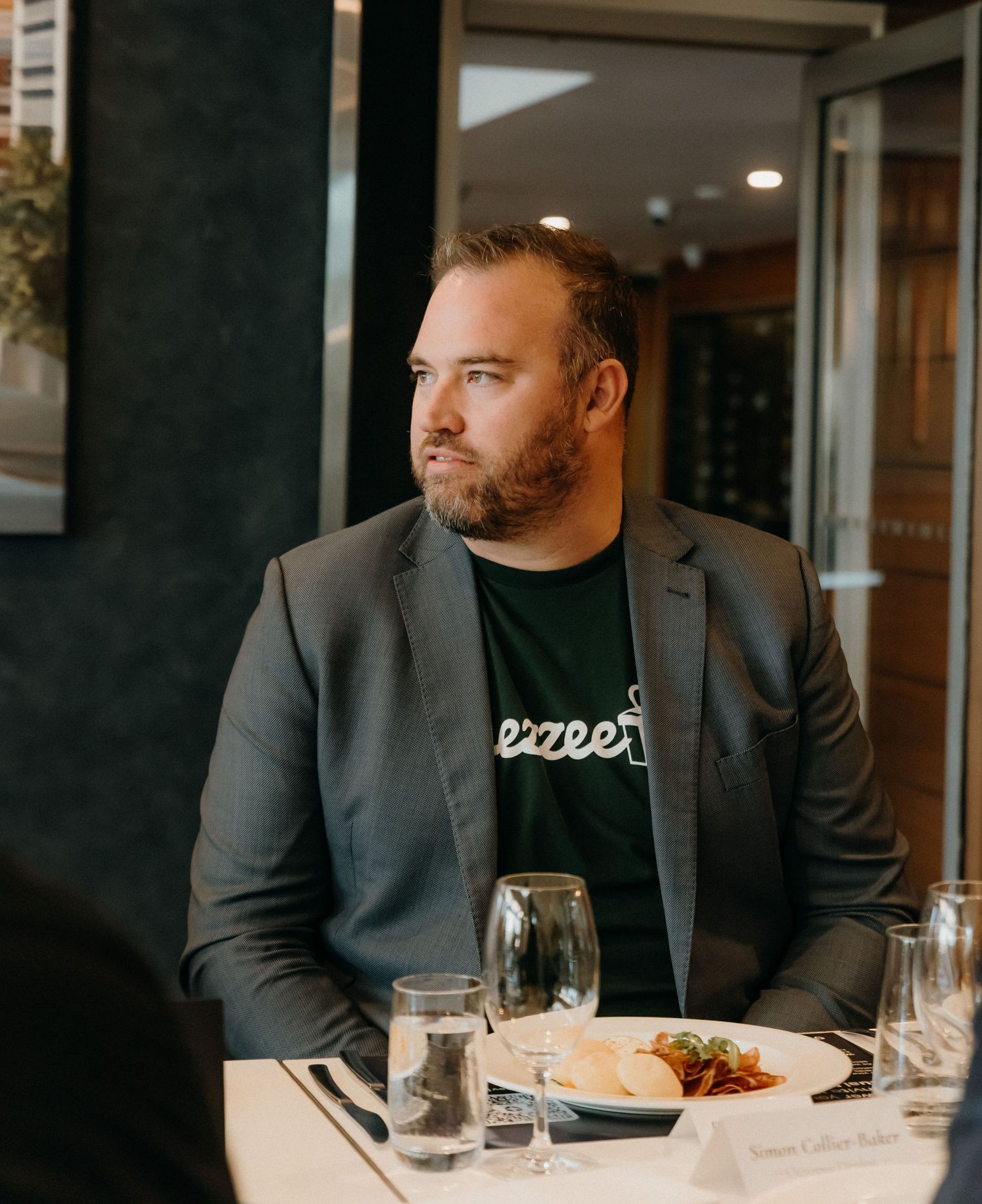 A person in a dark suit sits at a restaurant table with a plated meal and wine glass in front of them.