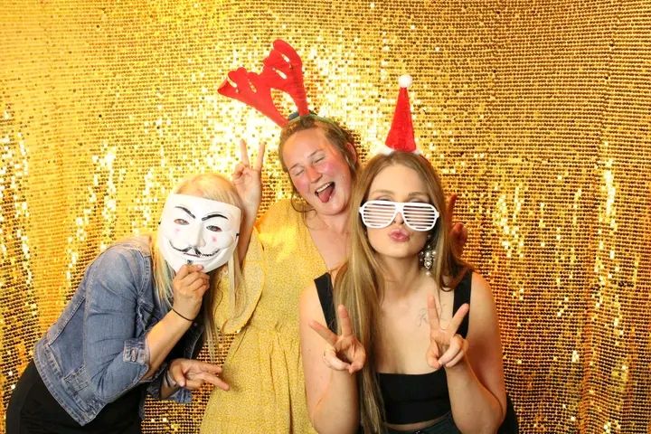 Three friends pose together with party props against a sparkly gold sequin backdrop at a photo booth.