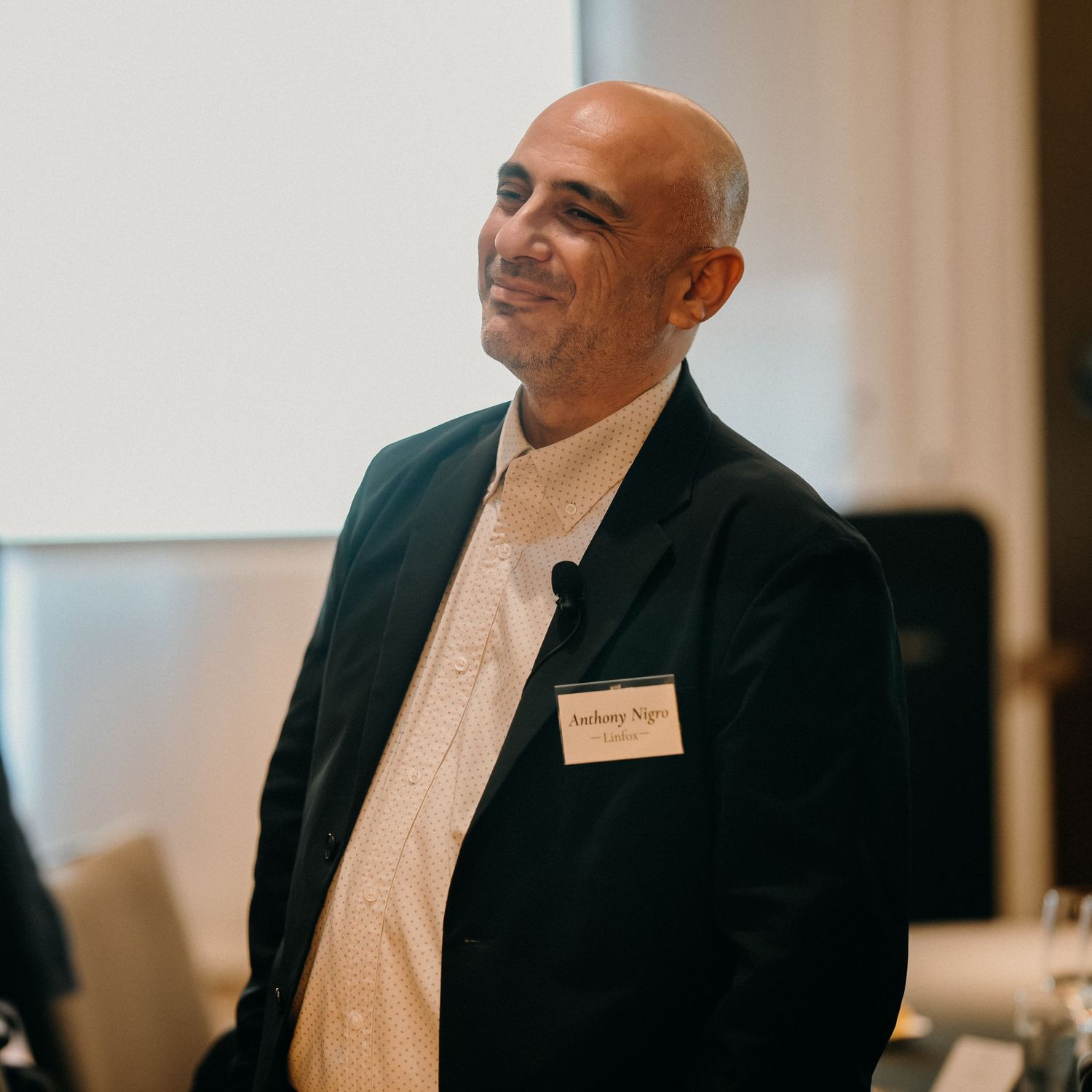 Person in a dark suit and white shirt smiling and gesturing while speaking during a presentation.