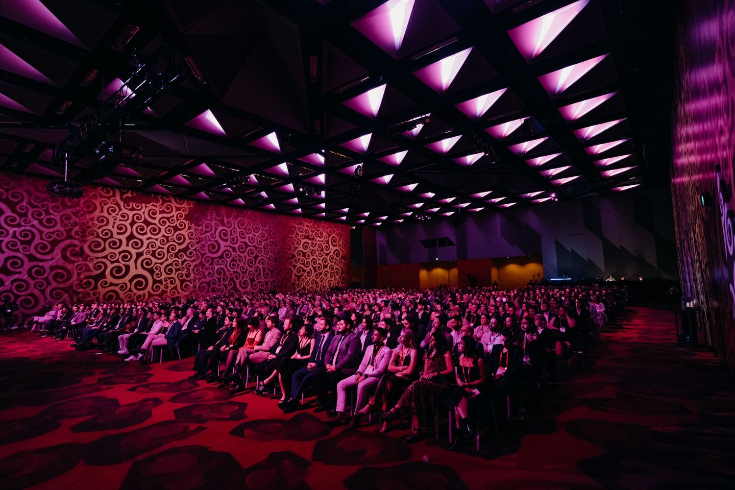Large audience seated in a dark auditorium with pink and purple lighting illuminating the space during an event.