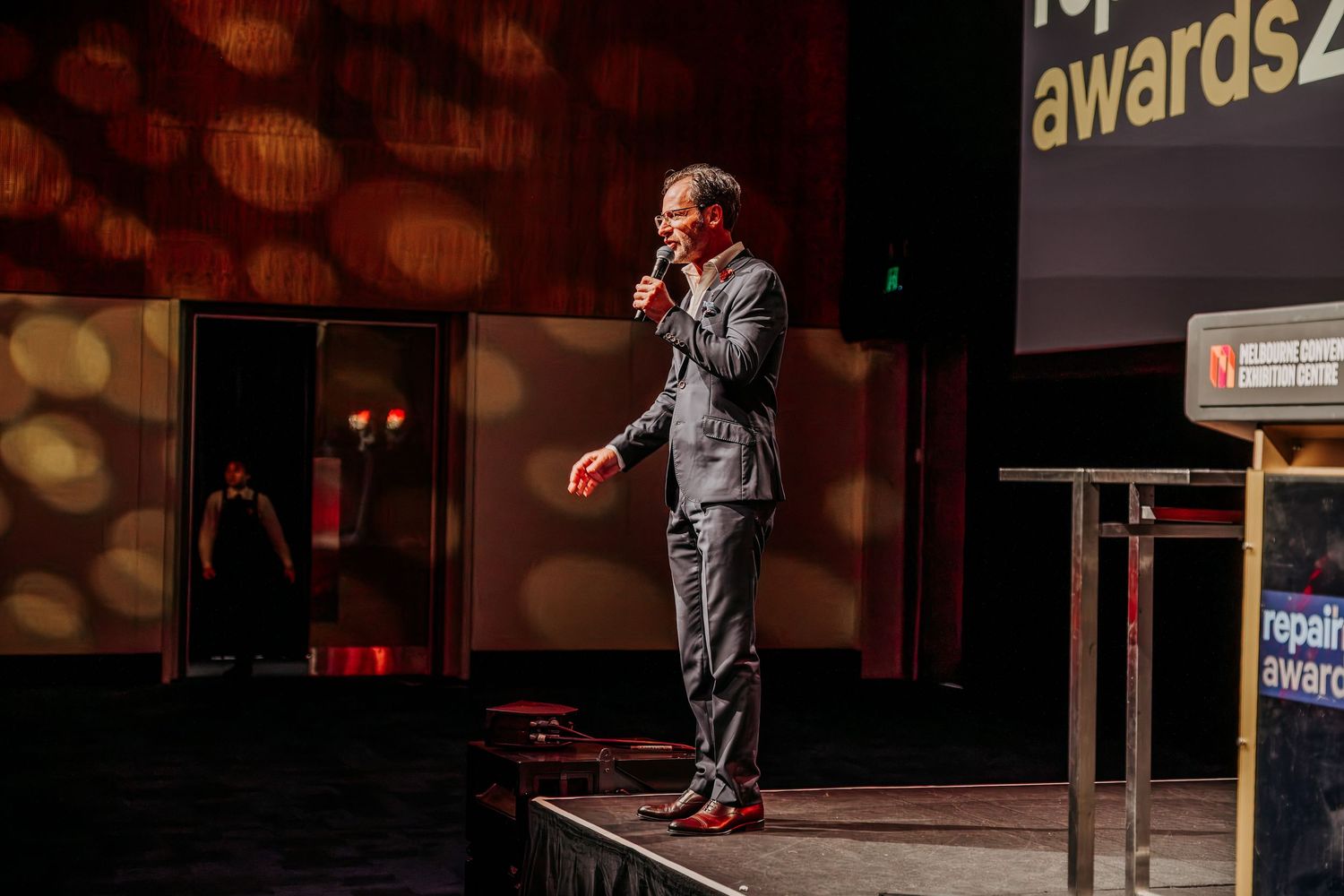 A performer in a gray suit stands on stage during a live comedy show with dramatic lighting and bokeh effects.