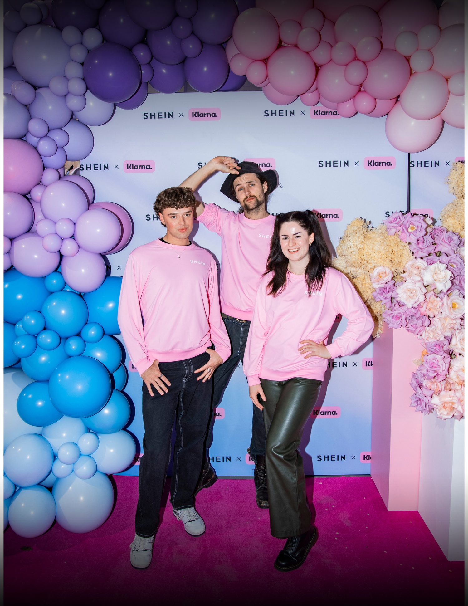 People in matching pink sweaters pose in front of a colorful balloon arch display with blue and pink gradient balloons.