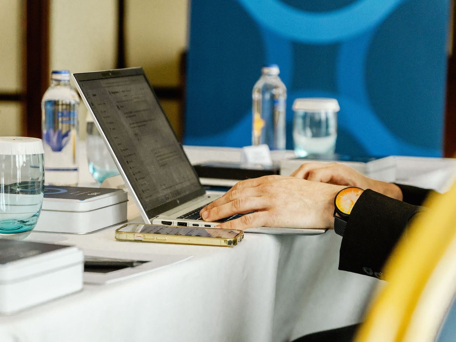 Professional typing on a laptop at a conference table with water bottles and notebooks in a modern meeting room.