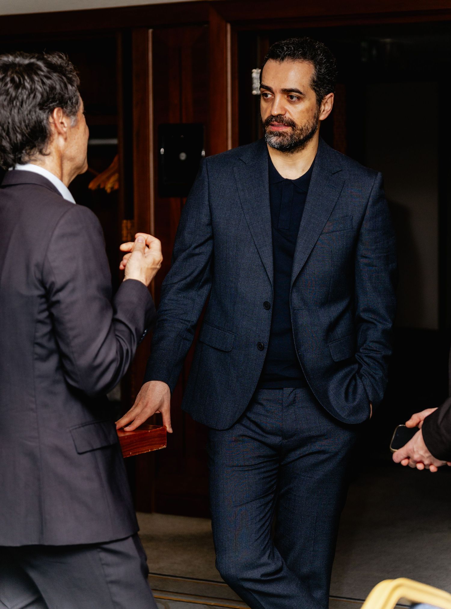 Two men in navy suits engaged in conversation near a doorway in a professional setting.