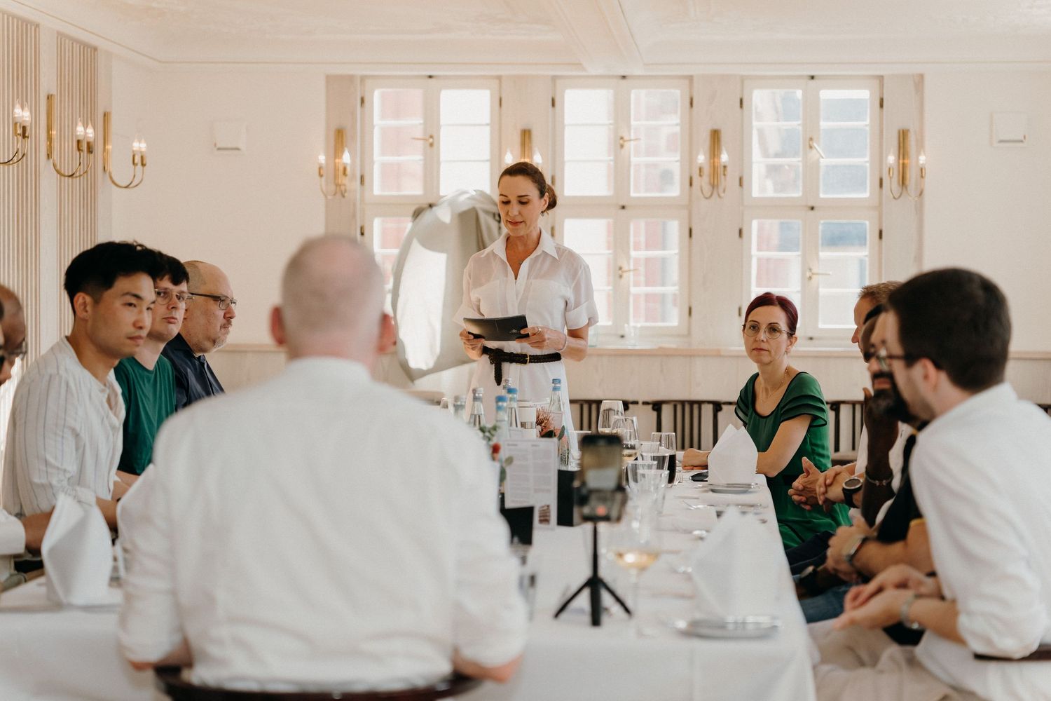 A waitress presents a dish to guests seated at elegant dining tables in a bright, upscale restaurant setting.
