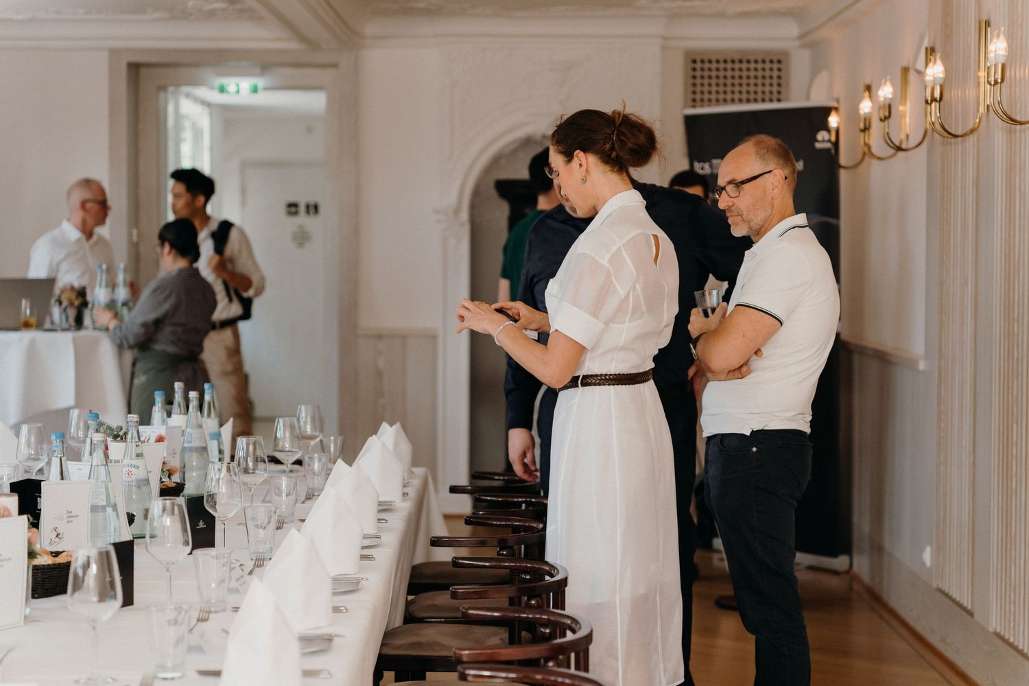 Elegantly set dining table with glassware in a formal banquet hall, guests mingling in the background.