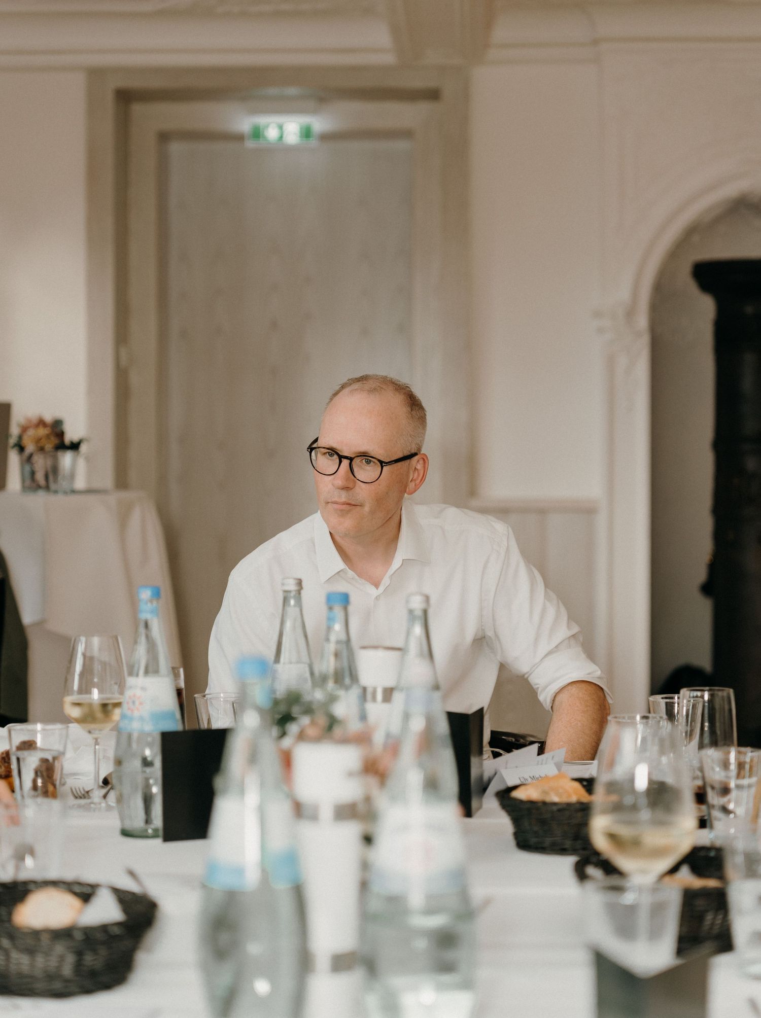 Man with glasses in white shirt sitting at an elegantly set dining table with water bottles and wine glasses.