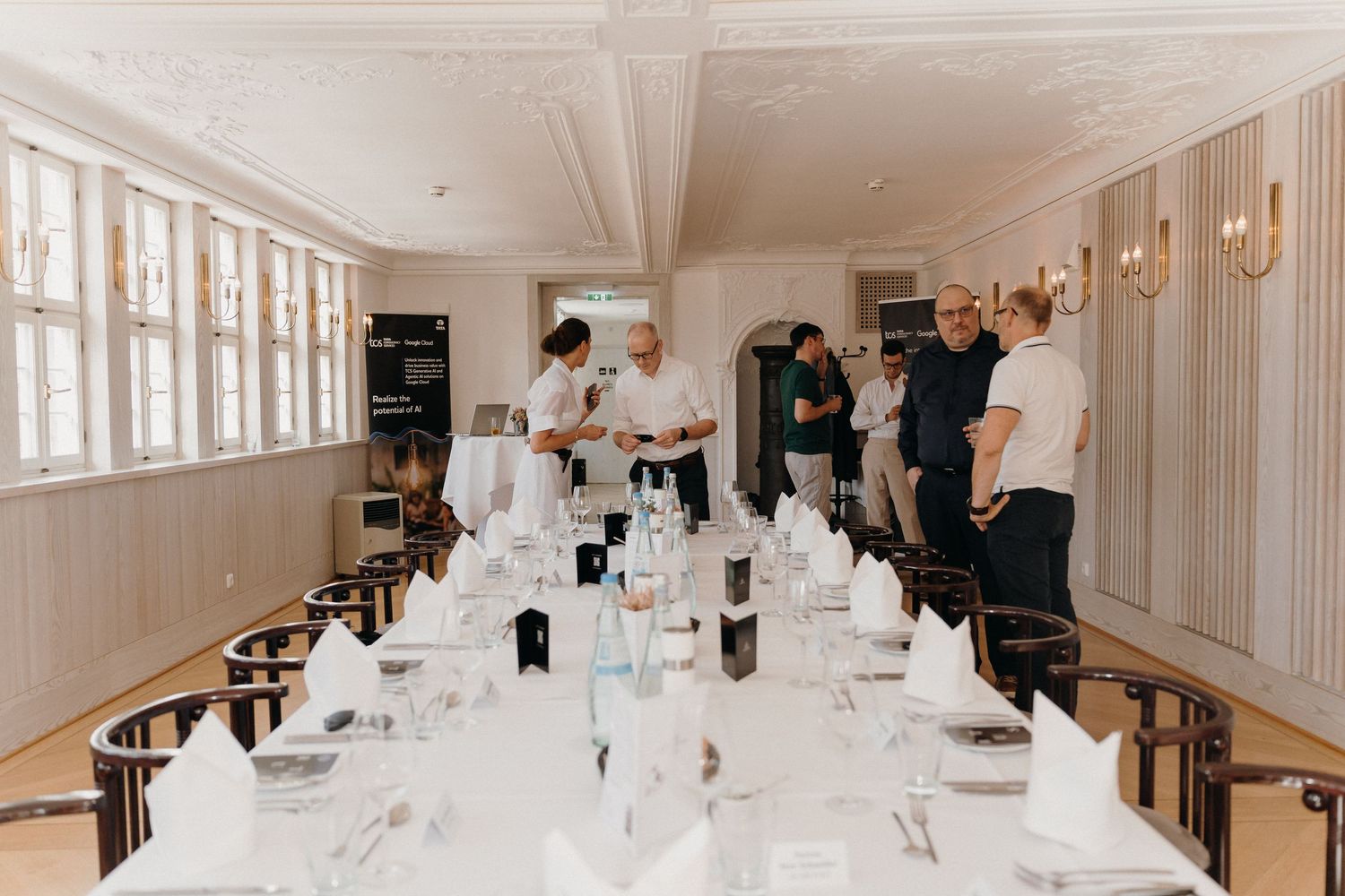 Elegant dining room with long white-clothed table set for a formal event, staff preparing while guests mingle nearby.