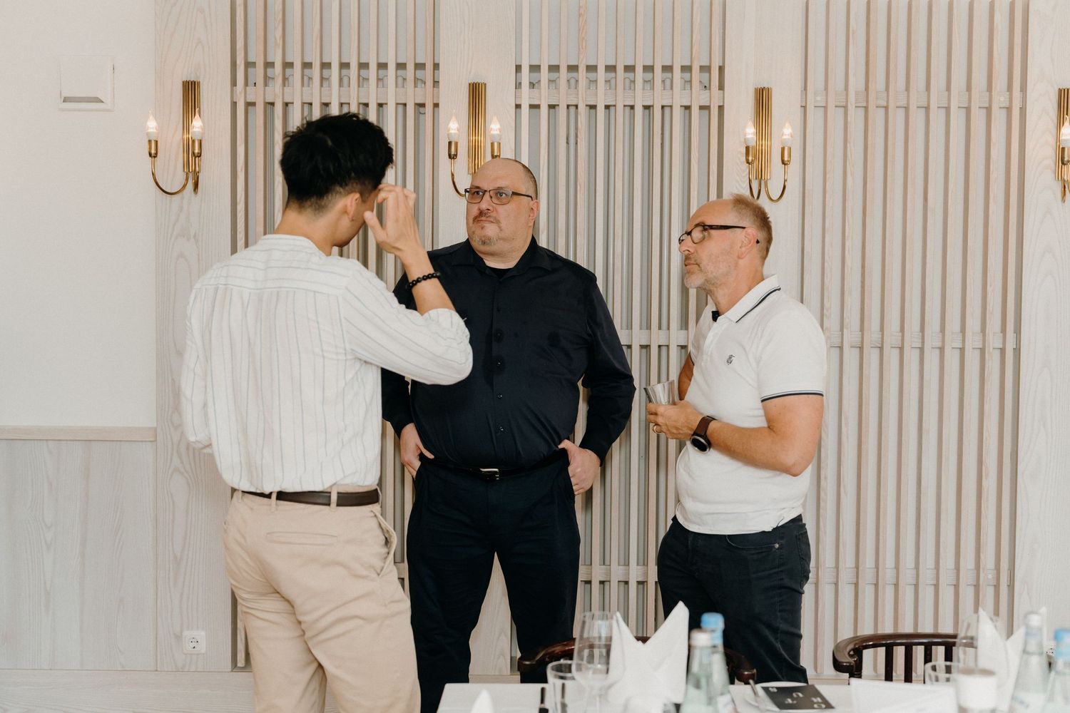Three men in smart casual attire engaged in conversation inside an elegant dining room with wall sconces.
