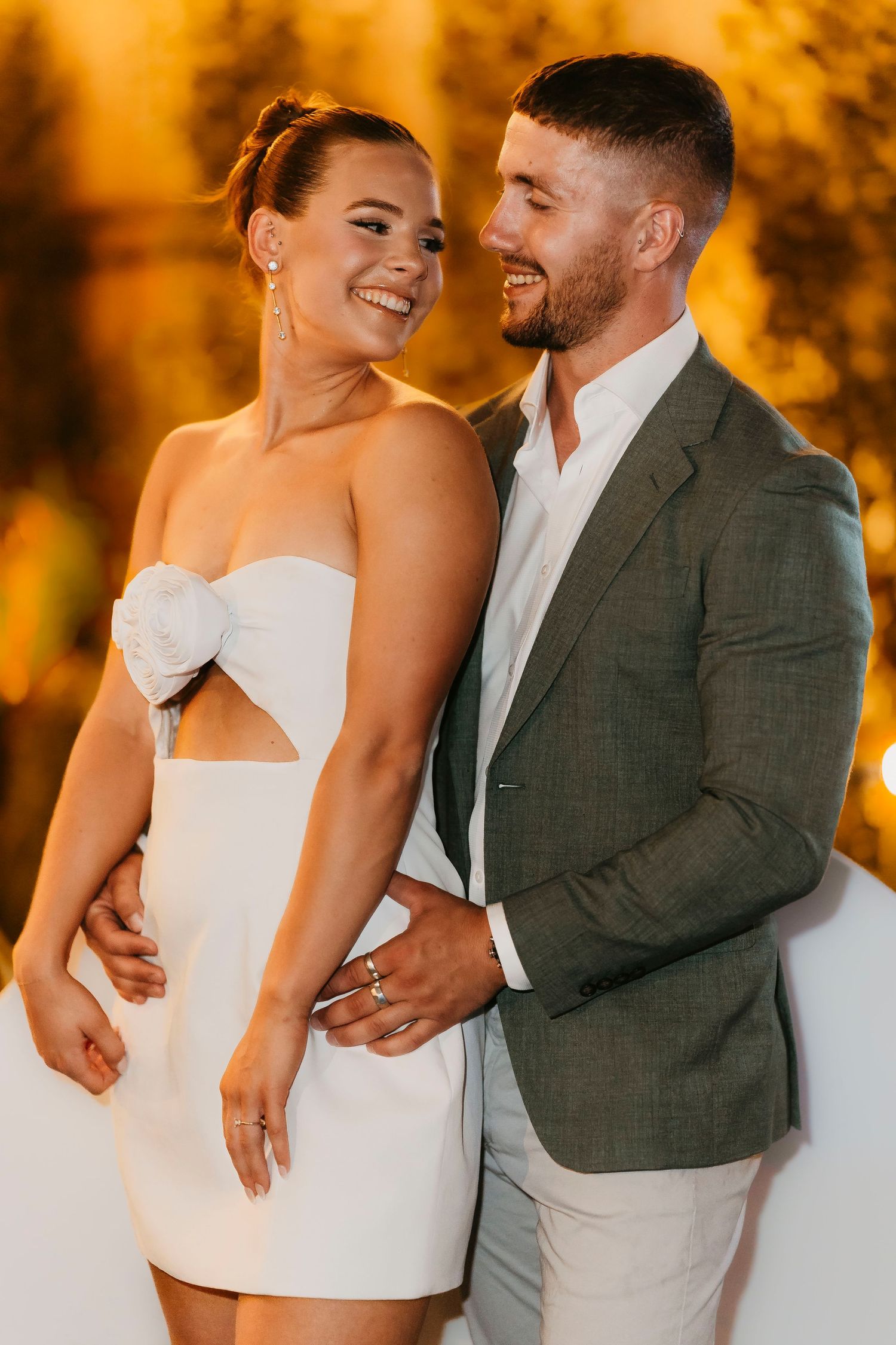 Couple embracing and smiling warmly at sunset wearing stylish formal attire with warm golden lighting in background.
