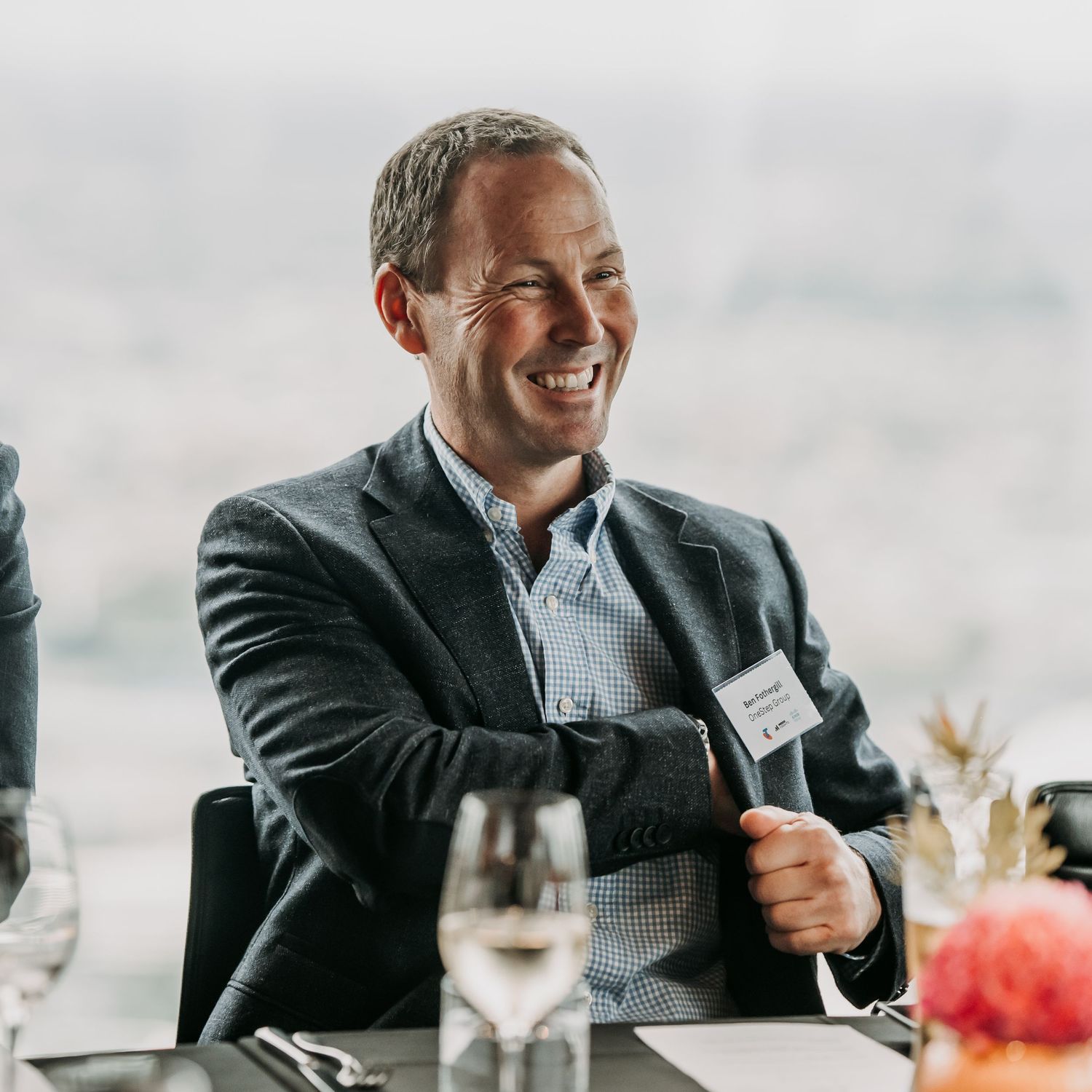 A person in a gray blazer and checkered shirt smiles warmly while seated at a formal dining table setting.