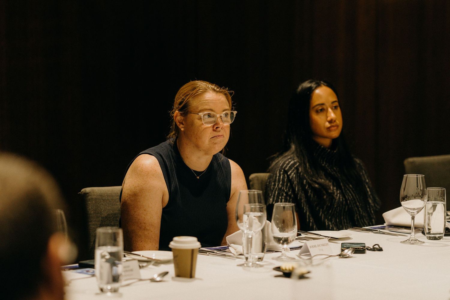 Two women seated at a formal dining table during a conference or business event with glassware and coffee cups.