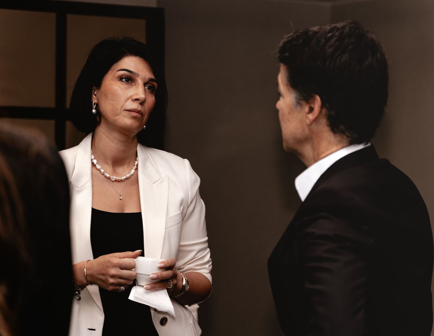 Two professionals in formal attire engaged in conversation, woman in white blazer holding a cup.