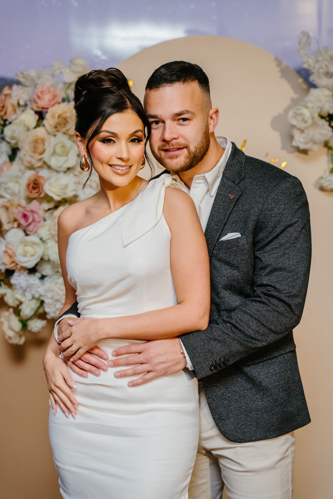 A couple poses together in formal wedding attire against a floral backdrop in a romantic photoshoot series.
