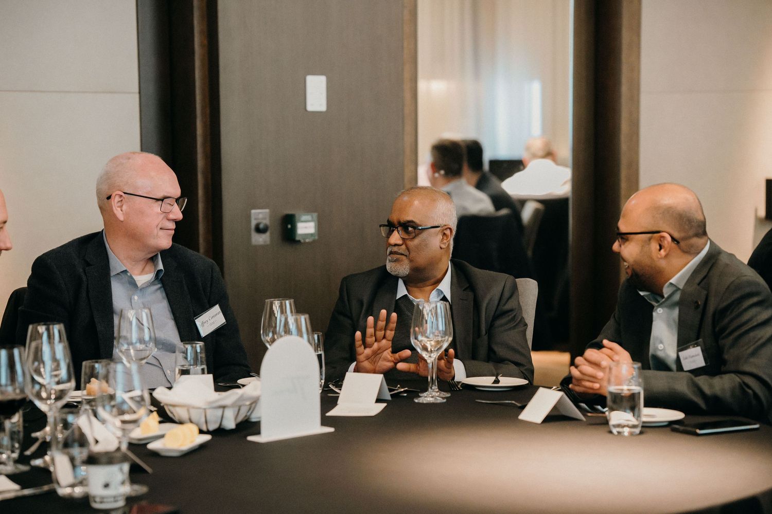 Business professionals engage in discussion around a conference table with wine glasses and meeting materials.