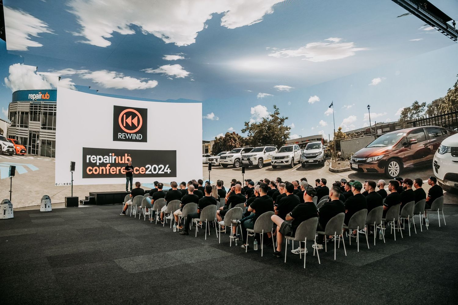 Outdoor event setup with rows of white chairs facing a stage under a blue sky with scattered clouds.