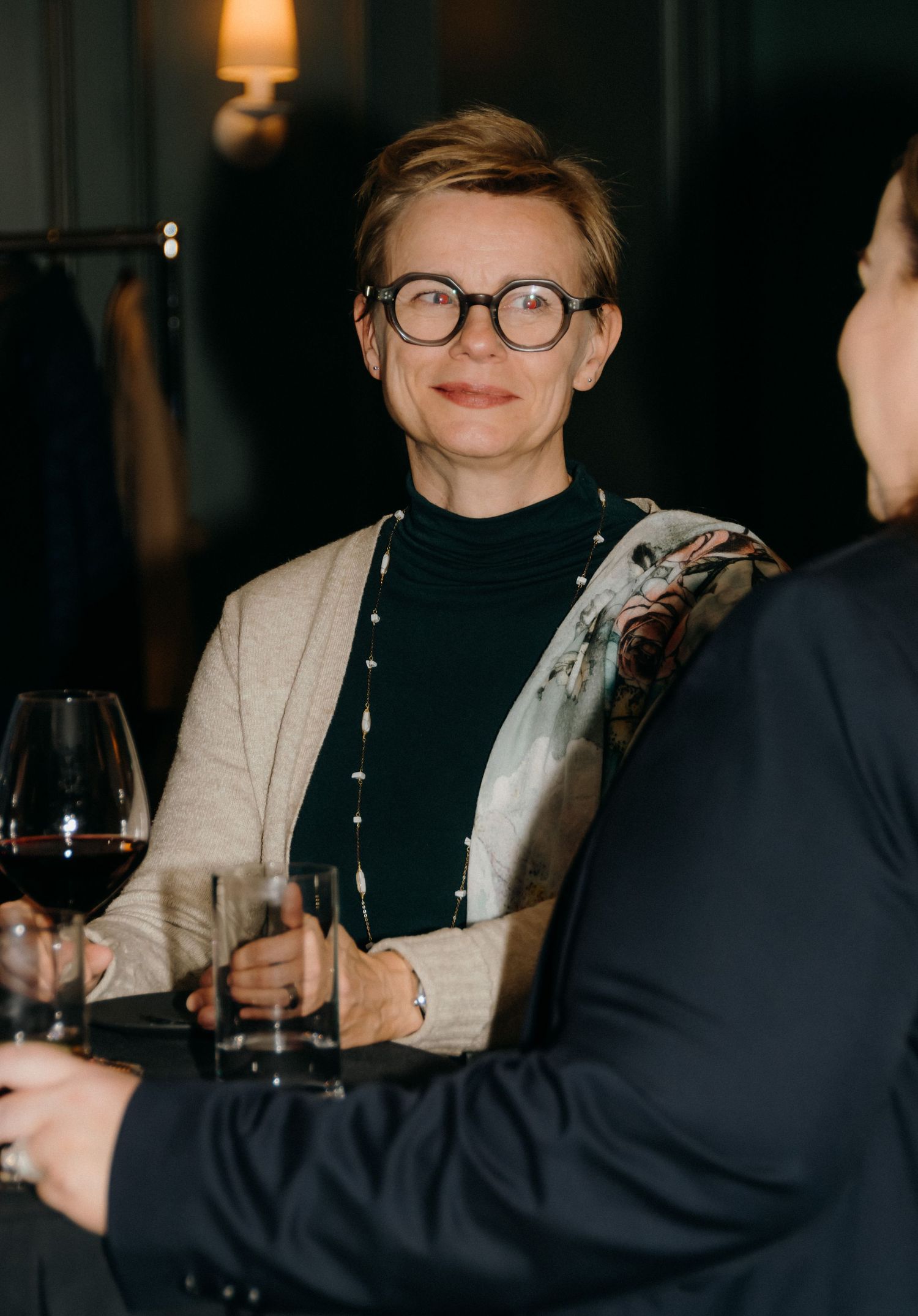 Two people in black attire share conversation over wine glasses in a dimly lit setting.