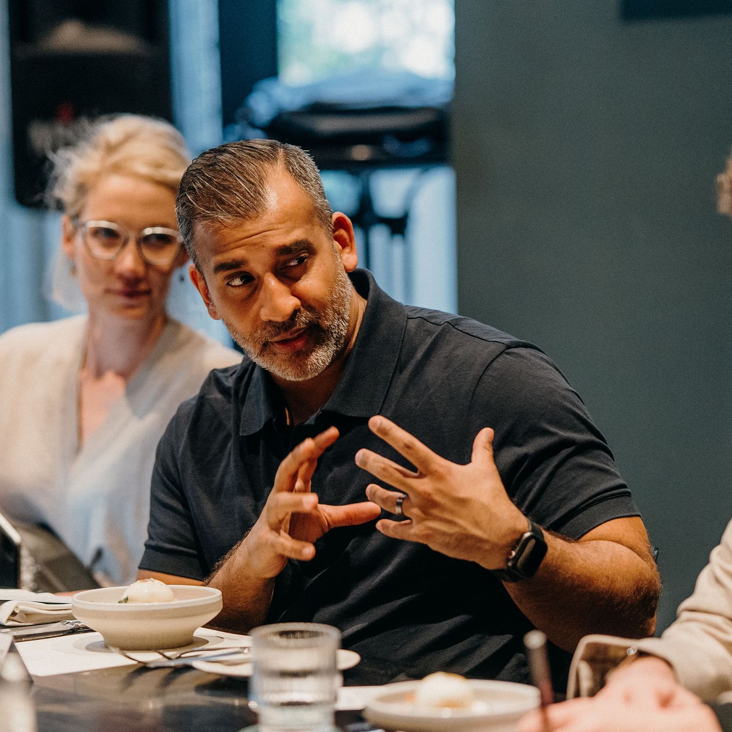 Person in black t-shirt gestures expressively during animated conversation at dining table.