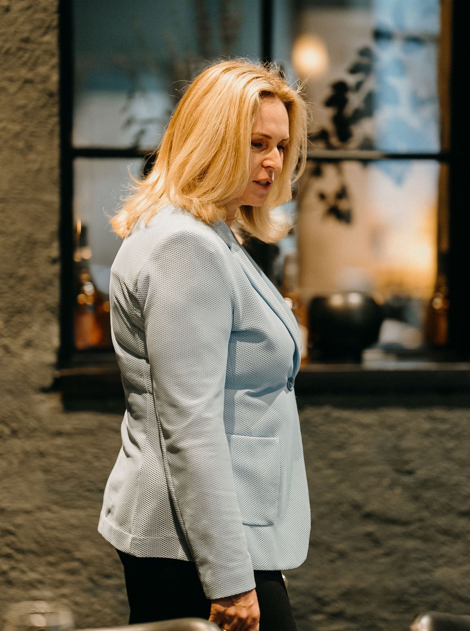 Woman in light gray blazer jacket poses against stone wall in moody evening lighting.