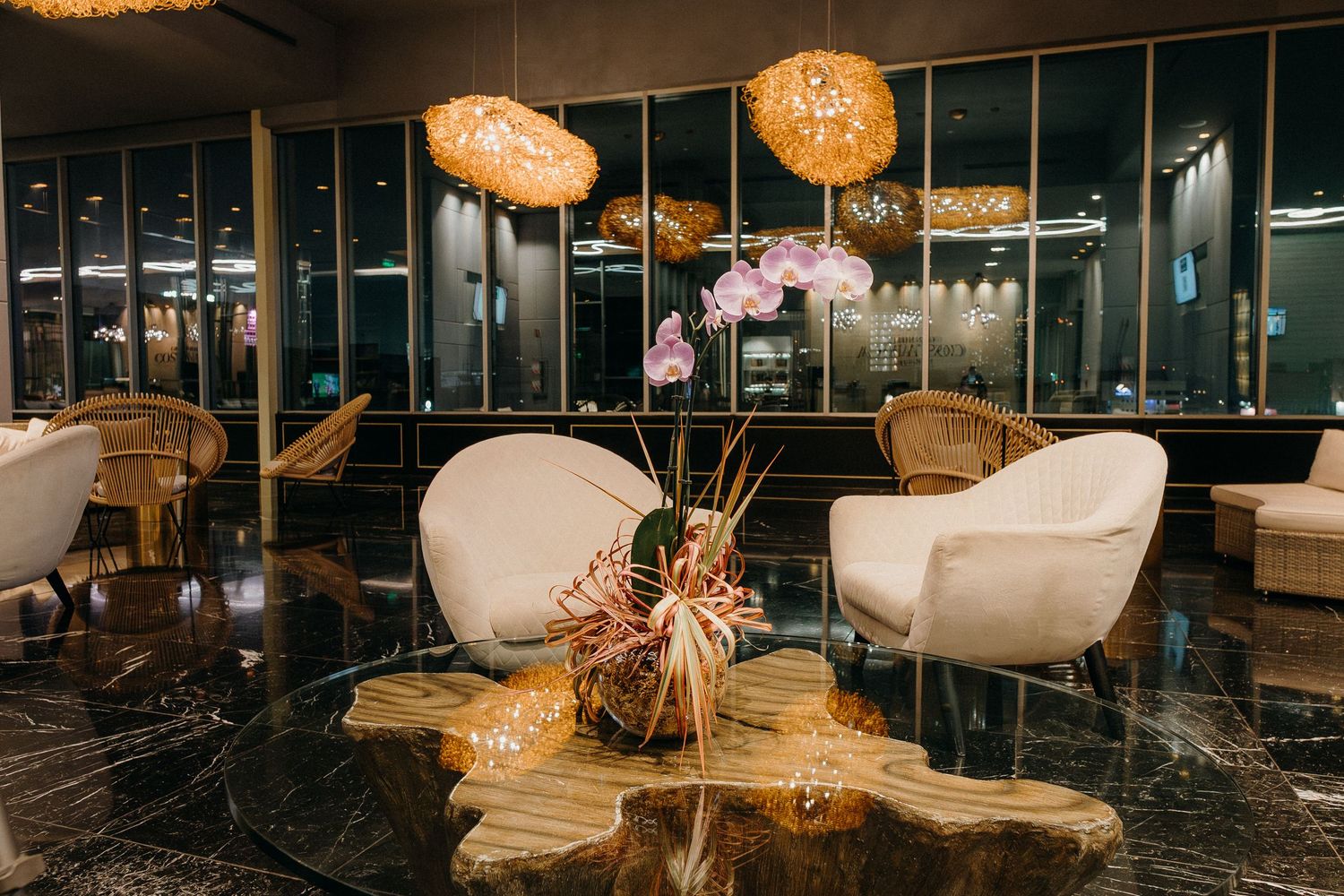 Modern hotel lobby with white curved chairs, woven pendant lights, and dark marble floors featuring a stylish floral centerpiece.