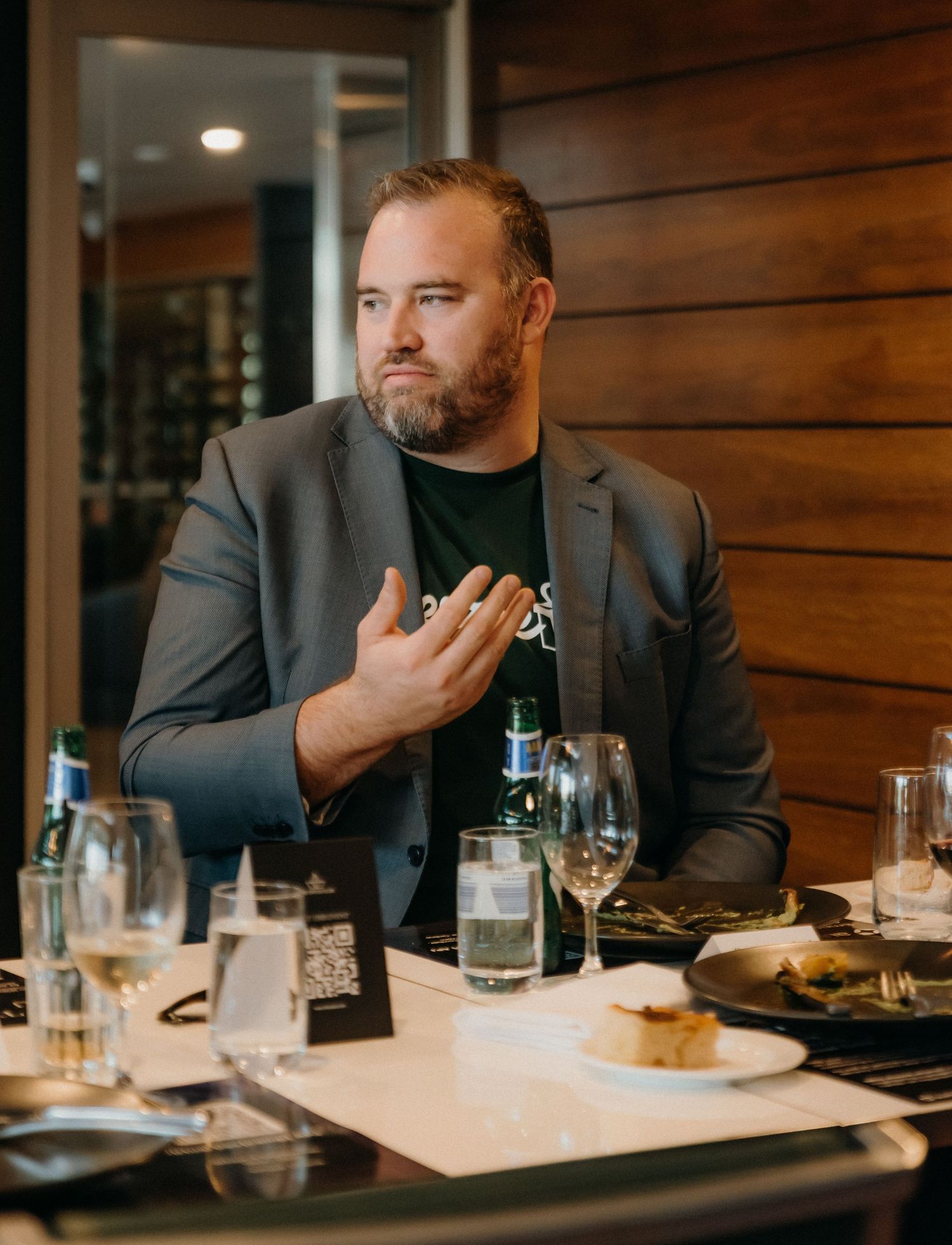 A person in a gray suit jacket sits at a restaurant table with glassware and white plates in warm lighting.
