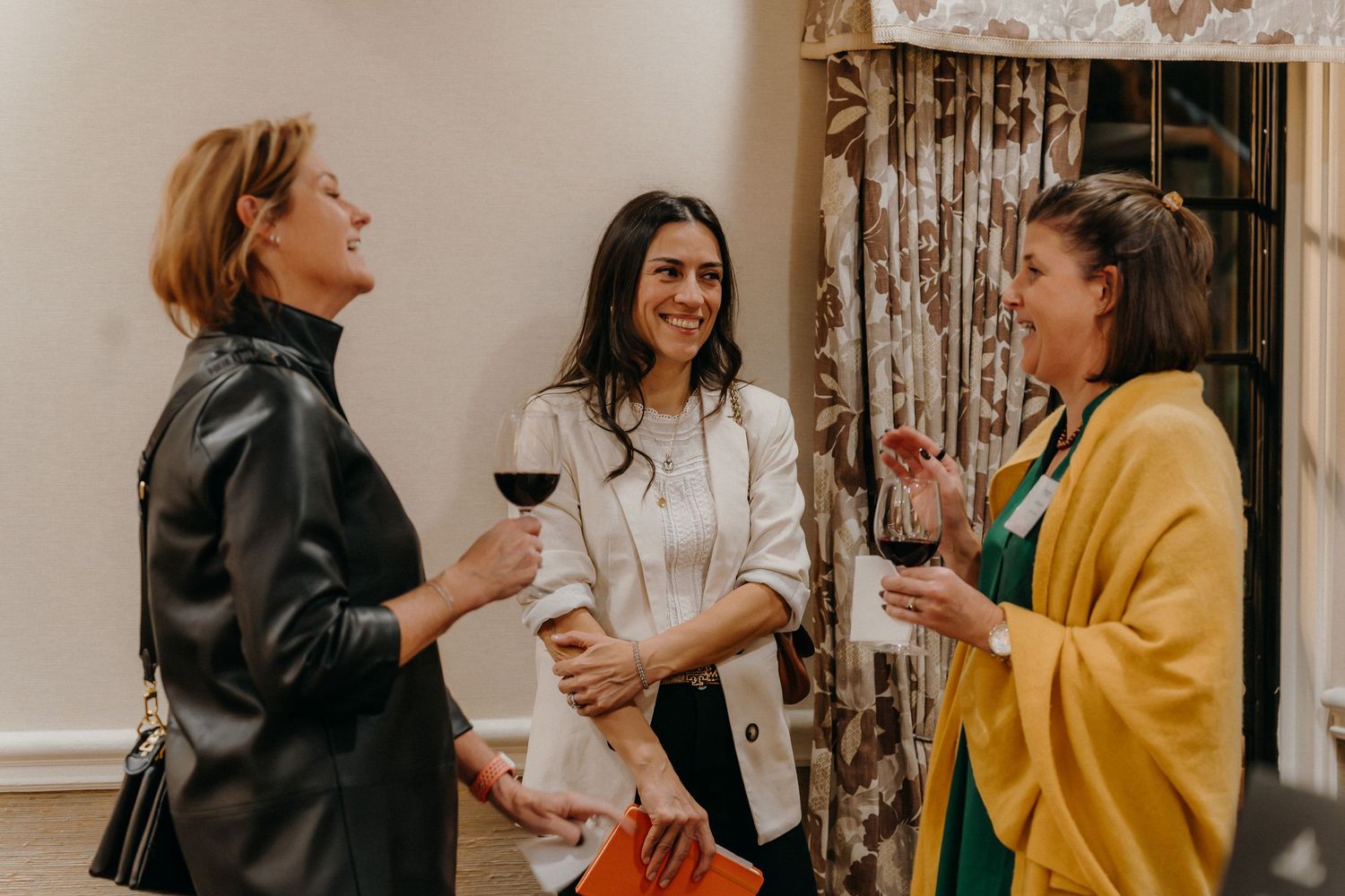 A group of women chat and laugh together while holding wine glasses at a social gathering.