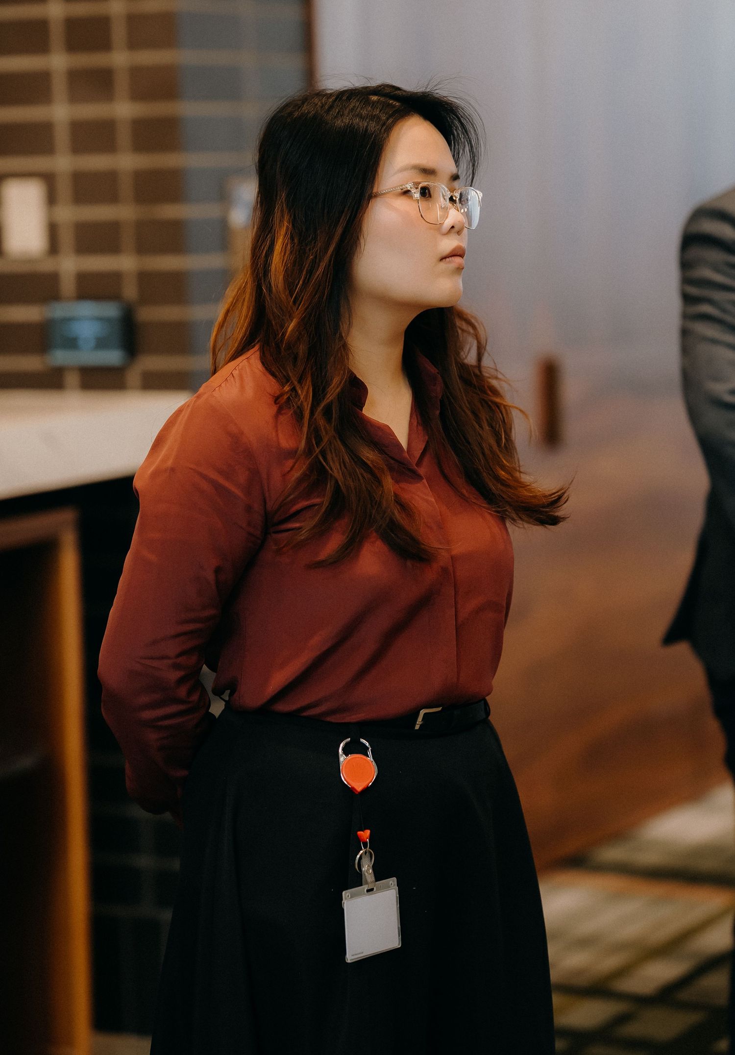 A person in a burgundy top and black skirt stands thoughtfully in an office setting with warm lighting.