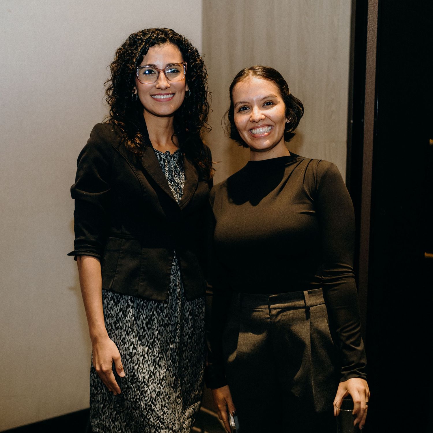 Two people posing together in professional attire against a light colored wall at an event.