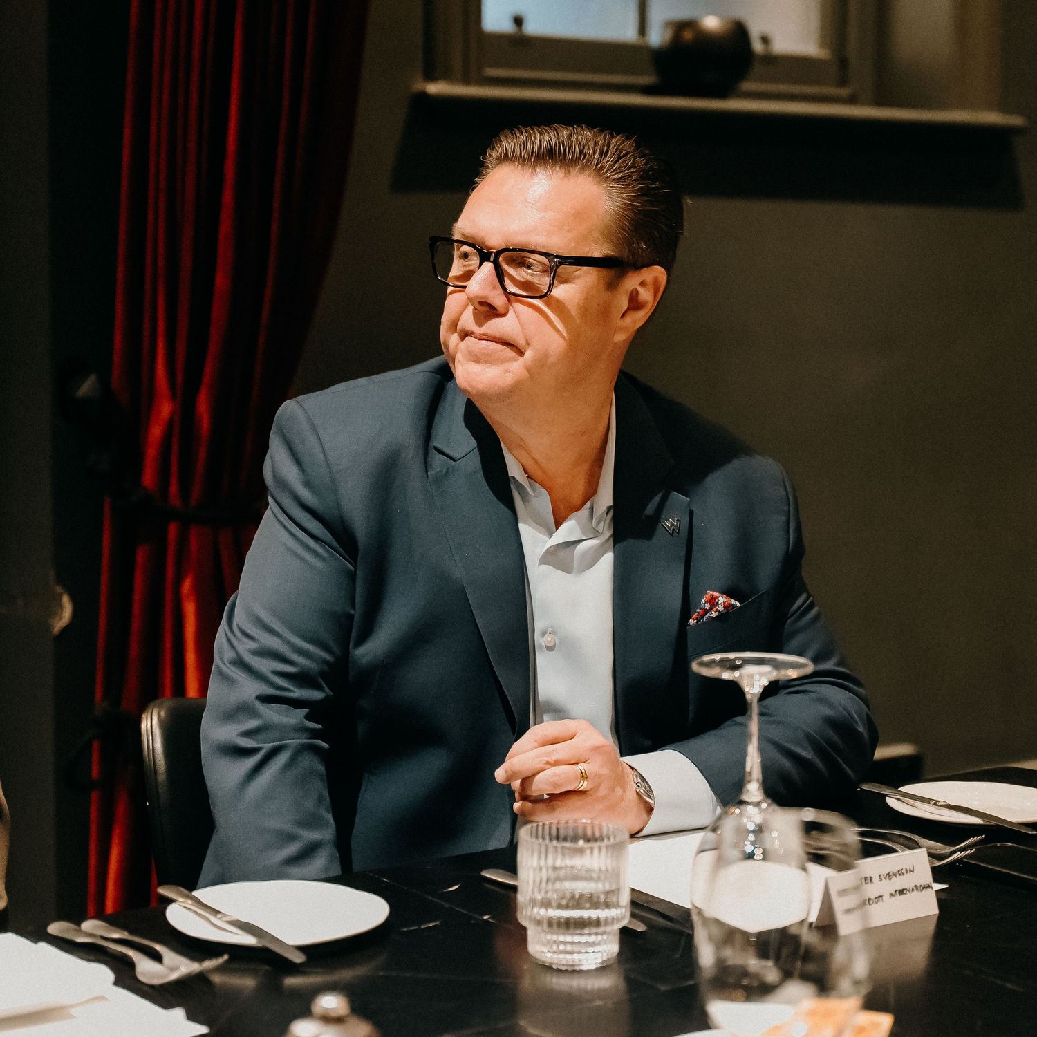 A person in a navy suit and white shirt sits at a restaurant table with glasses and plates.