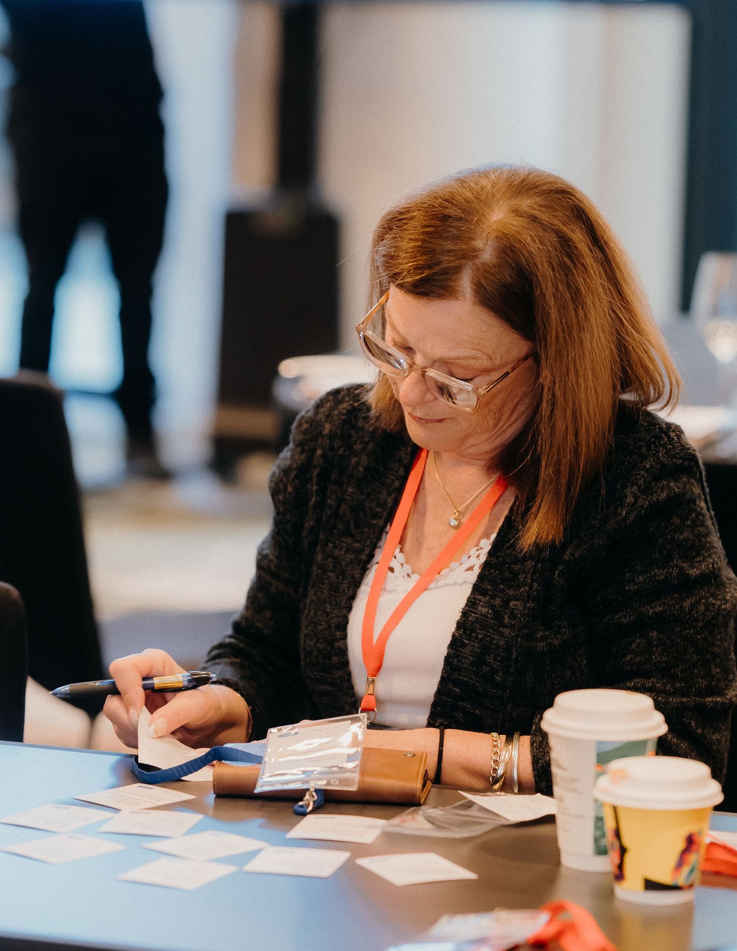 Professional wearing a black blazer writes notes at a conference table with a disposable coffee cup nearby.