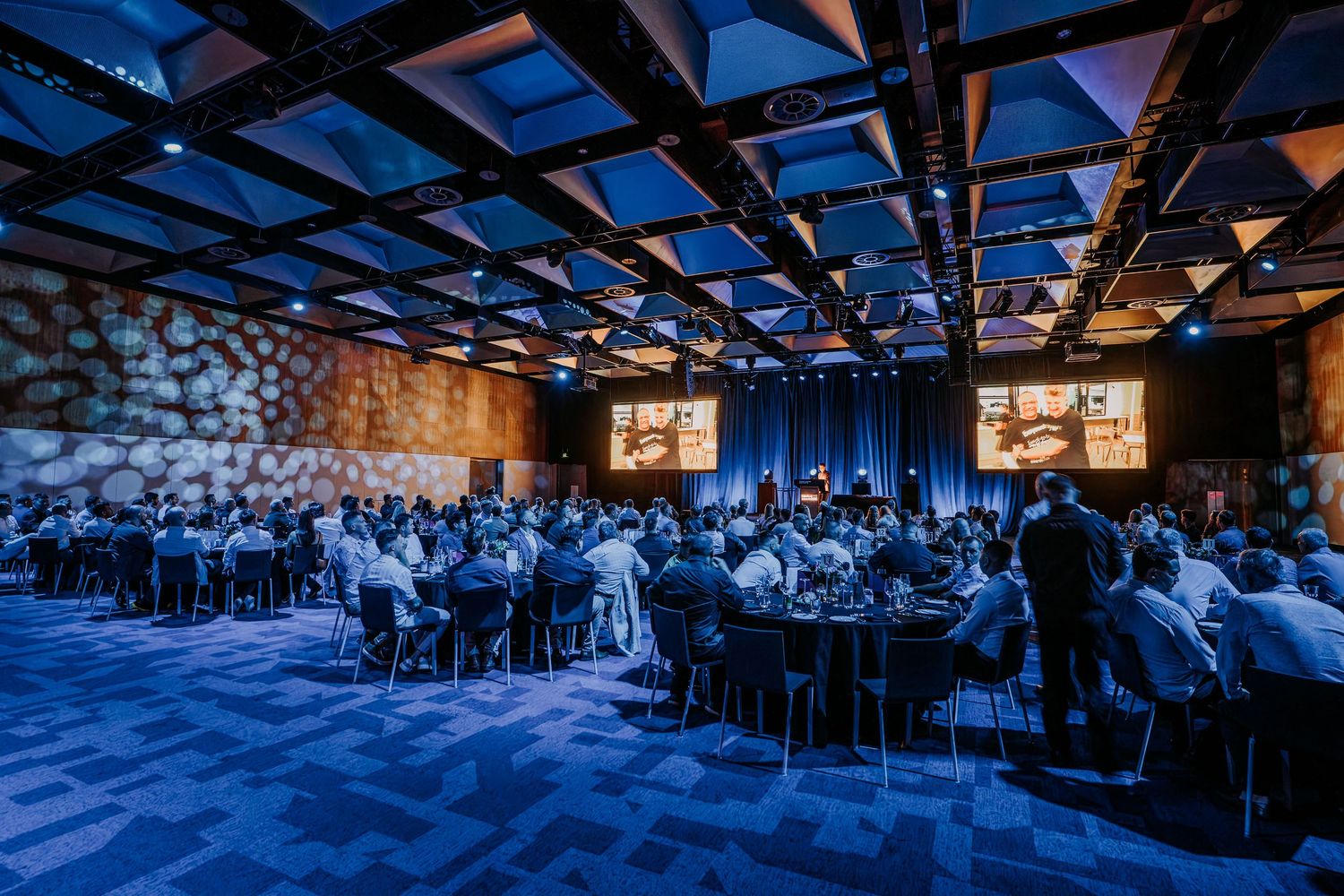 A panoramic view of a crowded conference hall with blue lighting and attendees seated in rows during a presentation.