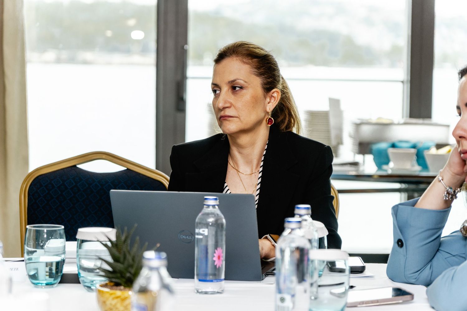 Professional woman in black blazer sitting at conference table with laptop and water bottles at business meeting.