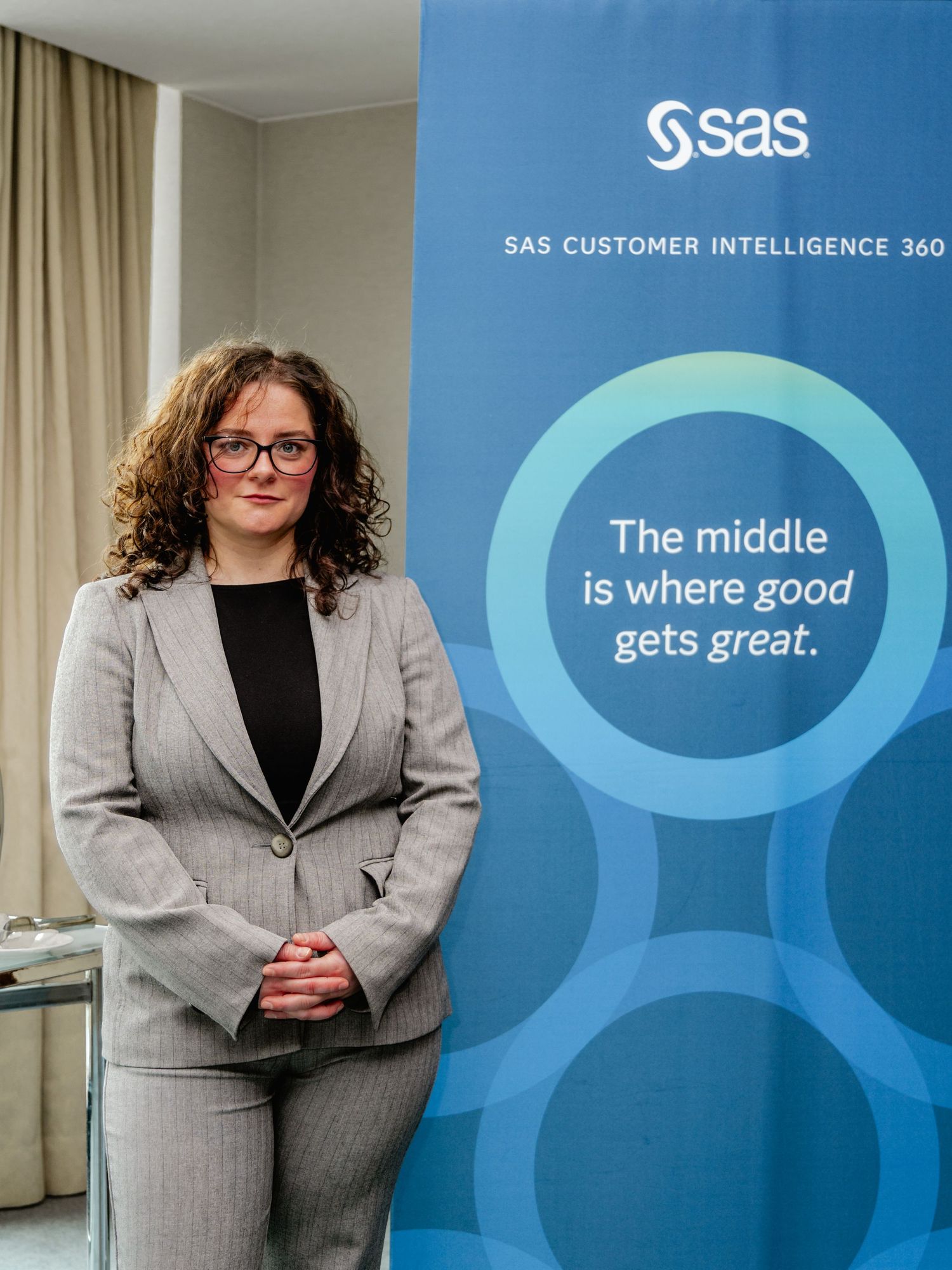 Professional woman in gray suit stands beside SAS Customer Intelligence 360 banner at corporate event.