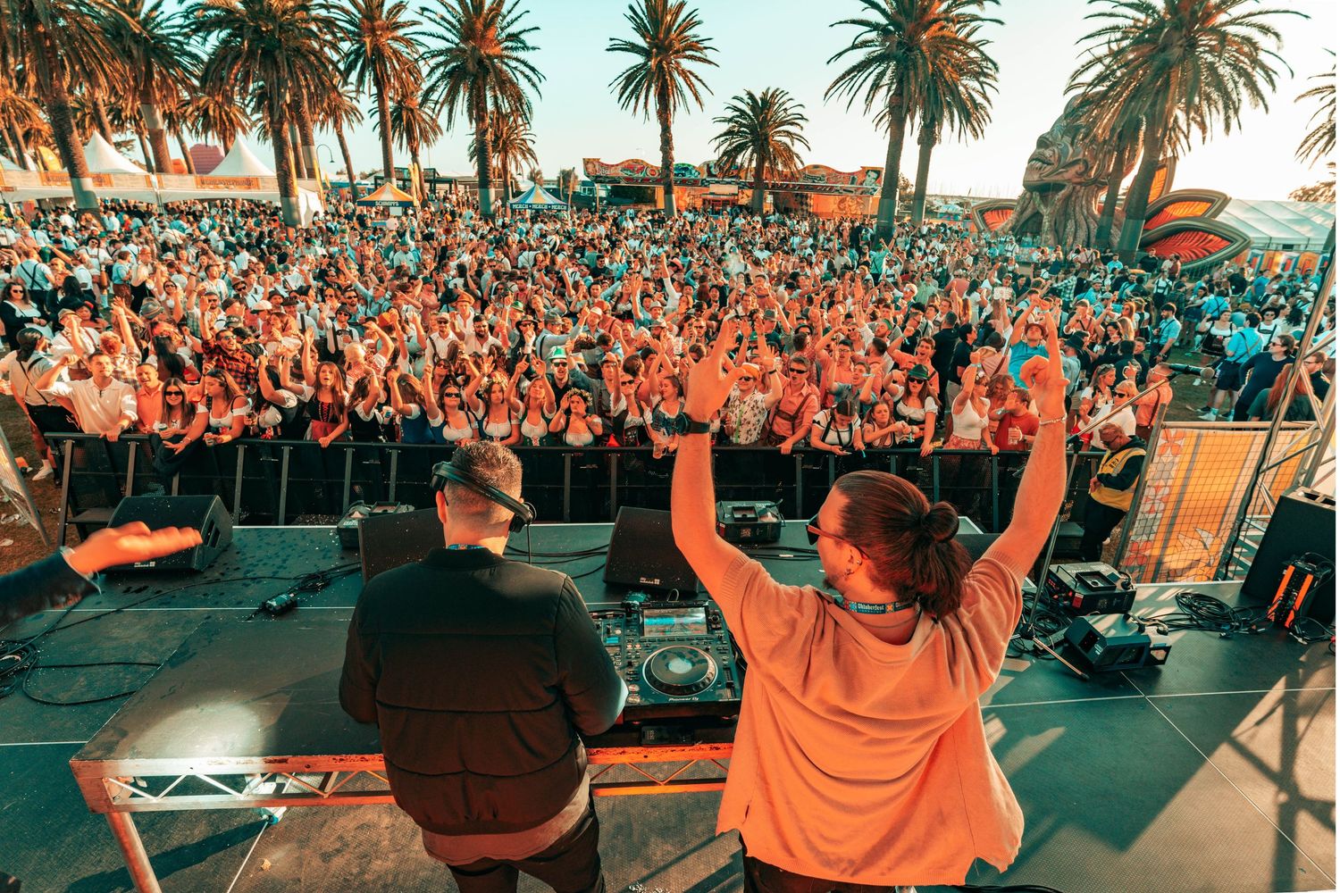 DJ performs at an outdoor music festival with large crowd and palm trees during sunset.