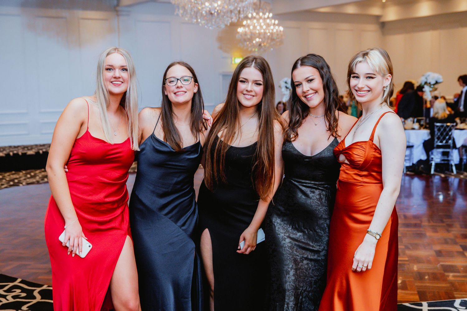 A group of friends pose together at a formal event wearing elegant floor-length dresses in red, black, and orange hues.
